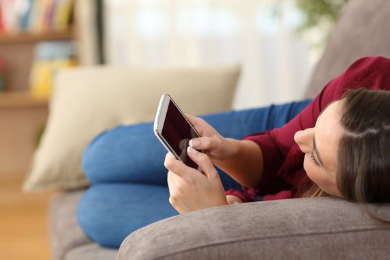 Une jeune femme étendue sur un sofa consulte son téléphone cellulaire.