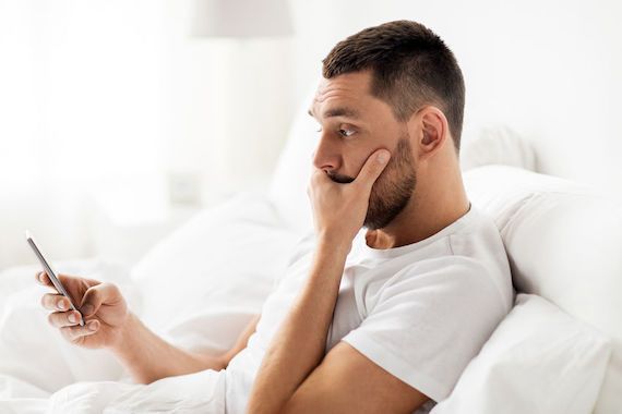 Un homme couché dans son lit regarde son téléphone cellulaire en étant surpris.